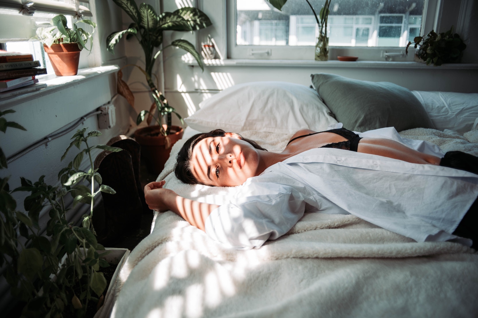 female laying in a white bed. she has on an open white shirt showing black underwear and there are plants around her.